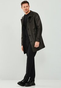 Zwarte leren jas met een gestructureerde afwerking, voorzien van een kraag, knoopsluiting en twee voorzakken. Gecombineerd met zwarte broek en schoenen.