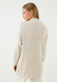 Sweter dzianinowy w jasnym beżu, o luźnym kroju, prążkowanej fakturze i długich rękawach. Widok z tyłu ukazuje subtelną kołnierz i opadające szwy ramion.