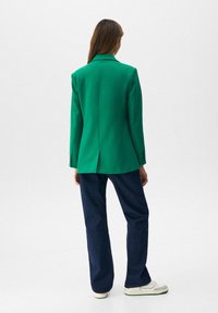 Blazer vert au design ajusté, avec une fente centrale, associé à un jean évasé bleu foncé et des baskets blanches.