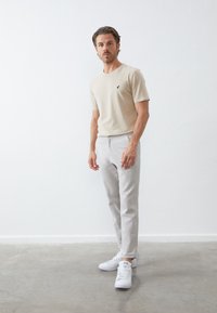 Camiseta de algodón beige clara con un pequeño logo negro, combinada con pantalones grises claros ajustados y zapatillas blancas. Textura suave.