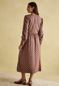 Ein langes, florales Kleid in pastellrosa mit Puffärmeln, tailliertem Bund und vollem Rock. Kombiniert mit burgunderfarbenen, niederen Schuhen.