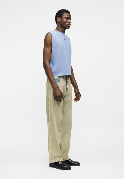 Άνδρας φορώντας αμάνικο ανοιχτό γαλάζιο πάνω μέρος, άνετο τζιν ανοιχτής πλύσης και μαύρα παπούτσια, στέκεται πλαγίως μπροστά σε λευκό φόντο.