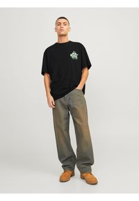 Černé oversized tričko s zeleným grafickým potiskem, v kombinaci s volnými, vybledlými džíny a hnědými botami. Neutrální pozadí.