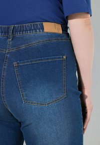 Jeans de mezclilla azul oscuro con una textura suave, que presentan una etiqueta de cuero marrón en la cinturilla y un único bolsillo trasero con costuras amarillas.