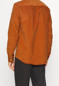 Camisa de pana marrón con un acabado texturizado, puños de botón y un diseño de dobladillo curvado. Cuenta con un yugo trasero para mayor estructura.