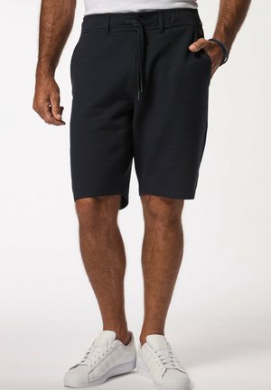 Homme portant un short noir texturé avec un cordon de serrage et des passants de ceinture, des baskets blanches et une chemise blanche rentrée, debout avec une main dans la poche.