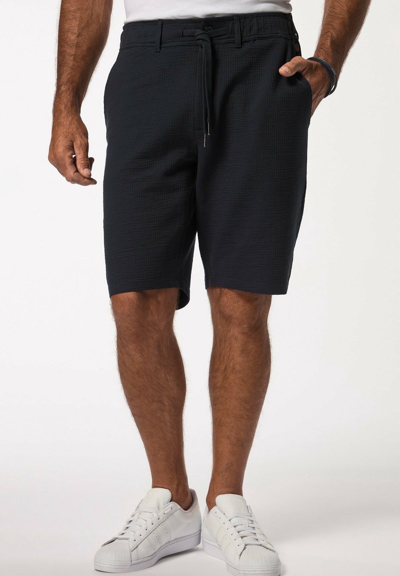 Homme portant un short noir texturé avec un cordon de serrage et des passants de ceinture, des baskets blanches et une chemise blanche rentrée, debout avec une main dans la poche.