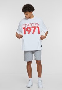 Camiseta blanca de gran tamaño con impresión roja "STARTER 1971", combinada con pantalones cortos grises y zapatillas blancas. Diseño sencillo y ajuste relajado.