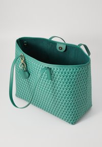 Teal schoudertas met een geometrisch patroon, textuurstof, twee handvatten en een charm accessoire. Binnenin heeft het een platte zak en een magnetische drukknoopsluiting.
