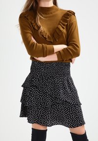 Brauner Pullover mit Rüschendetails am Ausschnitt, kombiniert mit einem schwarzen, gepunkteten, gestuften Rock. Das Outfit hat lange Ärmel und Kniehohe Stiefel.