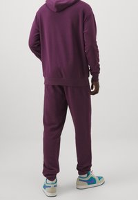 Fialová bavlněná mikina s kapucí v kombinaci s odpovídajícími tepláky, které mají žebrované manžety a volný střih. Tenisky s tyrkysovými a fialovými akcenty.