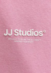 Lyserødt bomuldsstof med hævet hvid tekst, der lyder "JJ Studios" med mindre tekst nedenunder, der angiver mærkevedhæng og designtilhørsforhold.