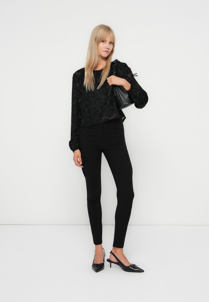 Czarna, teksturowana bluzka z długim rękawem w kwiatowe wzory, zestawiona z dopasowanymi czarnymi spodniami i szpilkami w szpic, uzupełniona małą czarną torebką.