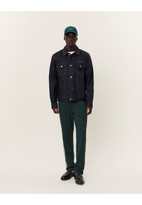 Giacca in denim scuro con due tasche sul petto e logo ricamato, abbinata a pantaloni sportivi verdi con strisce laterali e scarpe nere.