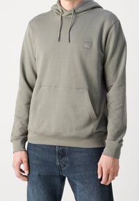 Homme portant un sweat à capuche gris clair avec une poche avant et un petit logo sur la poitrine, associé à un jean bleu, debout devant un fond uni.