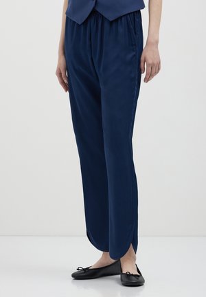 Persona che indossa pantaloni di satin blu navy con spacchi laterali e ballerine nere, in piedi contro uno sfondo chiaro e uniforme.