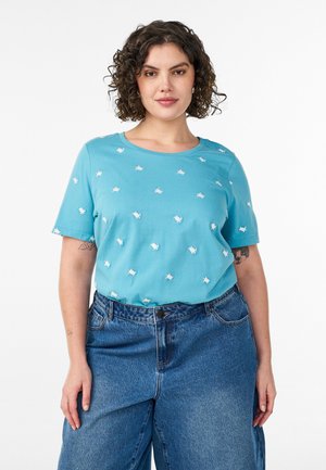 Kvinde med krøllet hår iført lyseblå kortærmet skjorte med mønster af små hvide skildpadder og løstsiddende blå jeans, stående mod en hvid baggrund.