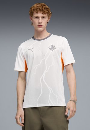 Jonge man met rood krullend haar, gekleed in een wit sportshirt met een grijze kraag, Puma-logo, bliksemschichtpatroon en oranje zijpanelen.