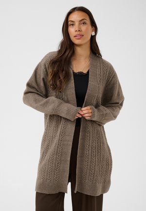 Femme portant un long cardigan marron en maille torsadée par-dessus un haut noir, debout les mains jointes et regardant devant elle sur un fond uni.