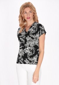 Top nero a stampa floreale con scollatura a V e maniche corte. Presenta un intricato disegno floreale bianco su uno sfondo nero. Abbinato a pantaloni bianchi.