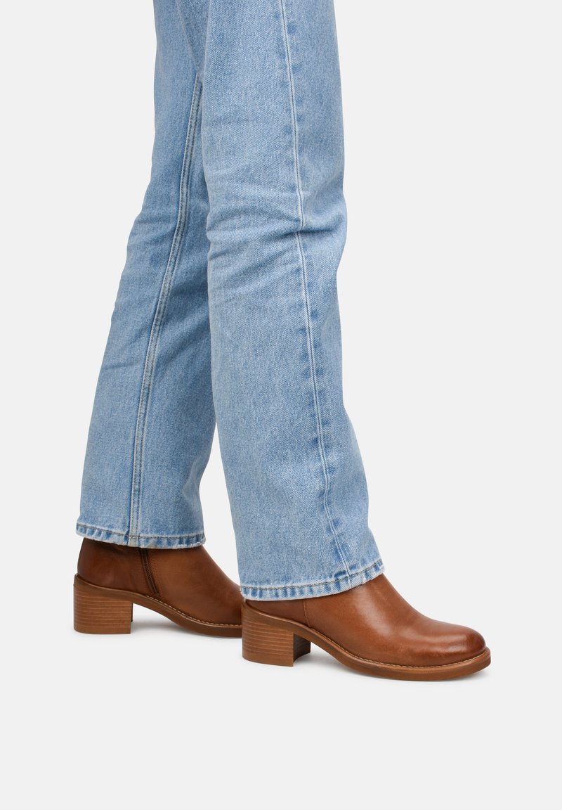 Botas tobilleras de cuero marrón con un tacón bajo y cuadrado. Textura suave, puntera redonda y detalle de costura mínima. Llevadas con vaqueros de color azul claro.