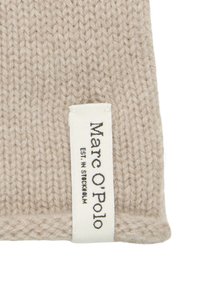 Beige gebreide stof met een getextureerd patroon, voorzien van een wit label met de tekst "Marc O'Polo" en "EST. IN STOCKHOLM" in zwart gedrukt.