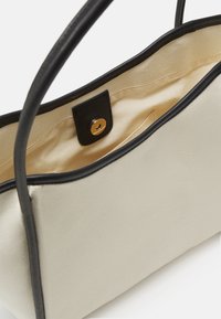 Beżowa torba z płótna z czarnymi skórzanymi akcentami, z zapięciem na złoty zatrzask i gładką podszewką. Długie czarne uchwyty do noszenia.