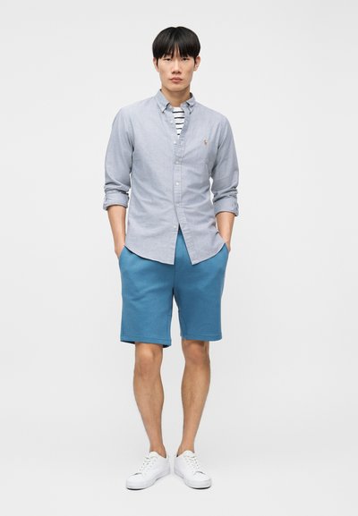 Άνδρας που φοράει ανοιχτό μπλε πουκάμισο με κουμπιά πάνω από ριγέ μπλουζάκι, μπλε σορτς και λευκά αθλητικά παπούτσια, στέκεται με τα χέρια στις τσέπες.