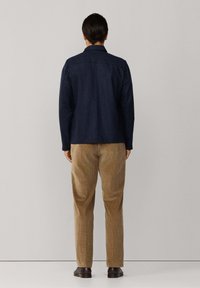 Donkerblauwe denimjacket met een gestructureerde kraag, gecombineerd met lichtbruine corduroy broek. De soepele textuur en de getailleerde pasvorm zijn duidelijk te zien.