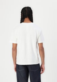 T-shirt de algodão branca com decote redondo e mangas curtas, apresentando uma bainha reta e um ajuste descontraído, combinada com jeans de denim azul escuro.