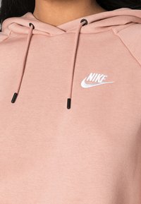 Roze hoodie met een zachte textuur, voorzien van een capuchon met trekkoord en zwarte koorduiteinden. Wit geborduurd Nike-logo op de borst.