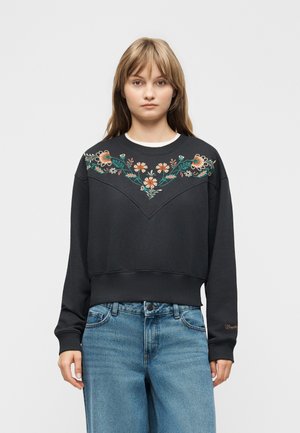 Young person wearing a black sweatshirt with colorful floral embroidery across the chest and light blue jeans, standing against a gray background.