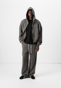 Grauer übergroßer Hoodie mit Kordeln, großen Taschen, kombiniert mit passenden lockeren Jogginghosen. Schwarzes T-Shirt darunter, flache schwarze Schuhe.