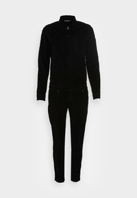 Combinaison en velours côtelé noir avec un devant à boutons, deux poches poitrine et des jambes fuselées, avec une texture lisse et une coupe ajustée.