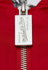 Roter Stoff mit einem Metallreißverschluss, der ein rechteckiges Etikett mit der Gravur "Mitchell & Ness" zeigt. Es hat silberne Töne und eine glatte Textur.