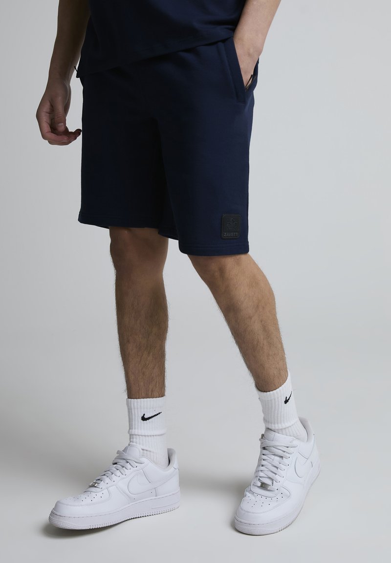 Homme portant un short bleu marine avec les mains dans les poches, chaussettes blanches Nike et baskets blanches Nike, debout sur un fond gris.