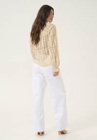 Beige lace blouse with long sleeves, featuring circular patterns. Paired with high-waisted white pants and beige heels. Light, airy texture.