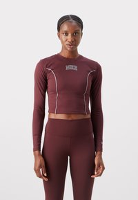 Viininpunainen pitkähiha crop toppi, joka on valmistettu joustavasta kankaasta. Siinä on valkoiset parisovitukset ja brodeerattu Nike-logo rinnassa.