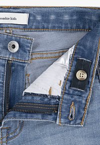 Jeans in denim con un lavaggio blu chiaro, caratterizzati da cuciture gialle a contrasto, patta con zip e chiusura a bottone con un accento nero vicino alla vita.