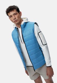 Gilet isolant bleu clair avec un design matelassé, fermeture éclair noire et poches, porté par-dessus un hoodie blanc avec cordon de serrage et un short beige.