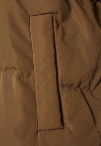 Tela de chaqueta acolchada marrón con una textura suave. Presenta un bolsillo de parche rectangular cosido a la superficie.