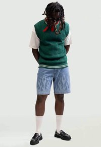 Green knitted vest over a white t-shirt, blue denim shorts with flame pattern, and black loafers. White crew socks complete the look.
