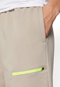 Beige sweatpants med en slät yta, med en liten ficka med blixtlås som framhävs av en neongrön dragkedja. Elastisk midja synlig.