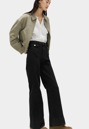 Kvinde iført en lys olivengrøn lynlåstrøje, hvid skjorte med knapper, sorte højtaljede viddebukser og brune mokkasinsko, stående sidelæns.