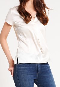 Donna che indossa una blusa bianca di raso a maniche corte con scollatura a V e jeans di denim blu, in piedi con una mano appoggiata all'anca.