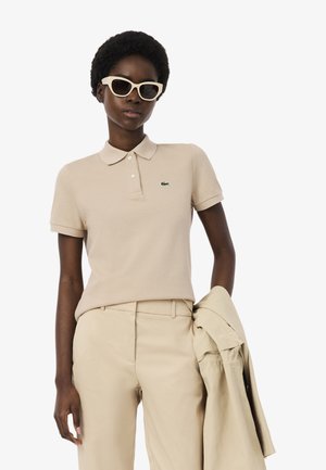 Frau trägt beige Polohemd, passende Hose und Cat-Eye-Sonnenbrille, hält eine beige Jacke in der Hand.