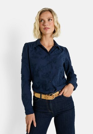Chemise boutonnière bleu marine avec broderies florales, manches longues et col classique. Associée à une ceinture beige et un jean foncé.