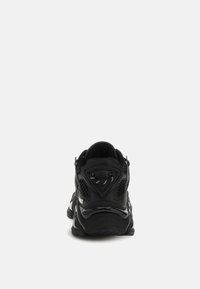 Musta urheilukenkä, jossa on teksturoitu päällinen, pehmustettu kaulus ja verkkokangasaksentteja. Kengässä on paksu pohja ja kolmionmuotoinen metalliyksityiskohta.