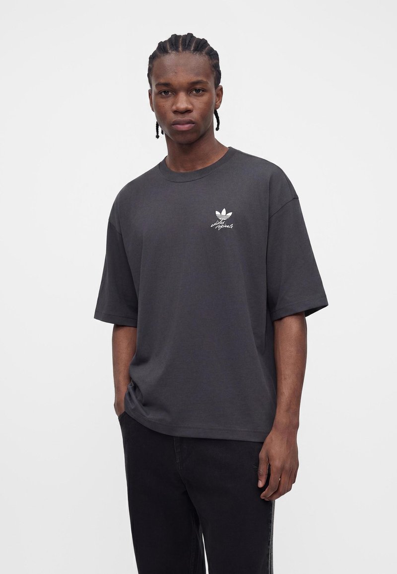 Junger Mann mit geflochtenem Haar, der ein dunkles, übergroßes Adidas Originals T-Shirt und schwarze Hosen trägt und vor einem schlichten hellen Hintergrund steht.