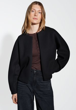 Veste noire courte avec des manches surdimensionnées, texture lisse et col rond, portée sur un haut métallique sombre et un jean noir.